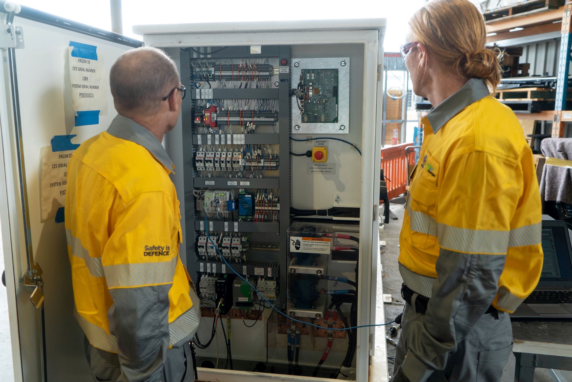 Two field crew standing in front of switchboard inside warehouse